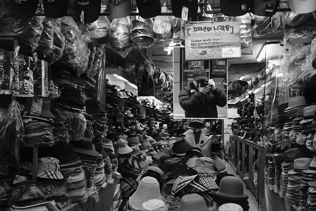 Black-and-white self-portrait of Craig Chee photographing his reflection in a mirror inside a hat shop crowded with stacked hats