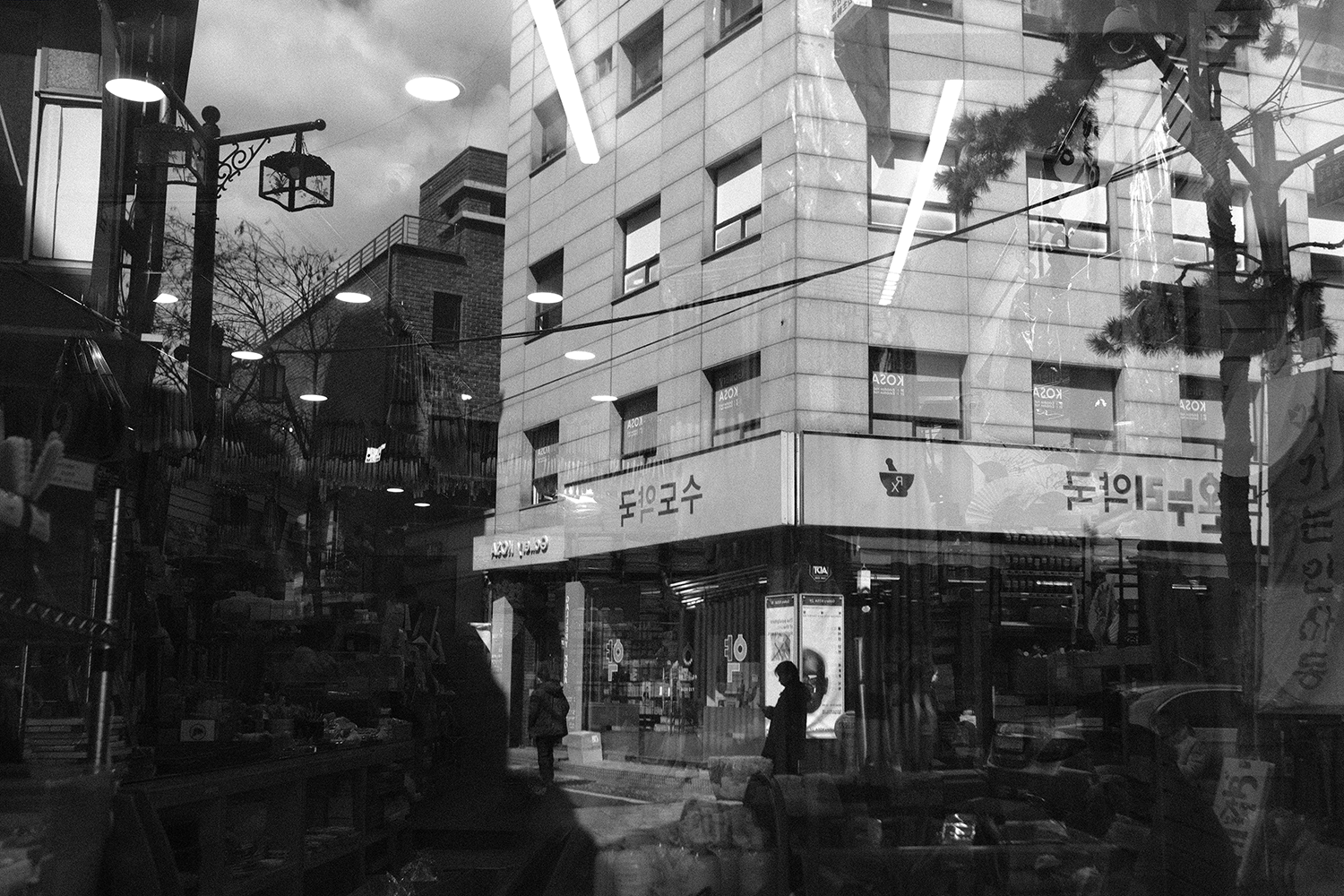 A street corner and pharmacy building seen through a shop window, its ceiling lights and hanging lanterns layered over the scene