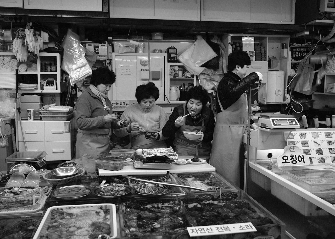 Four women in aprons sharing a meal behind the tanks of a crowded seafood market stall
