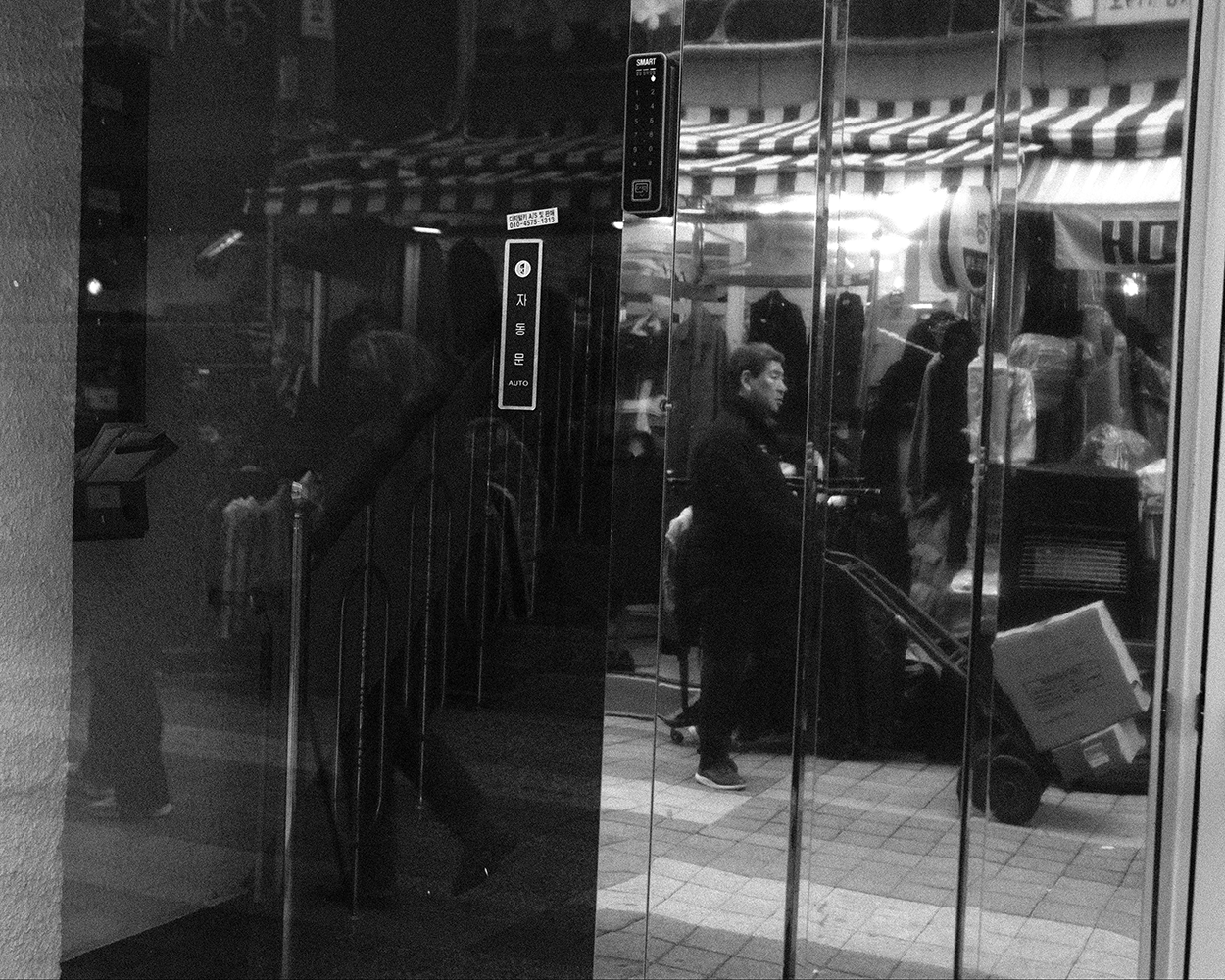 A man with a hand truck reflected in the glass doors of a building, a striped market awning behind him