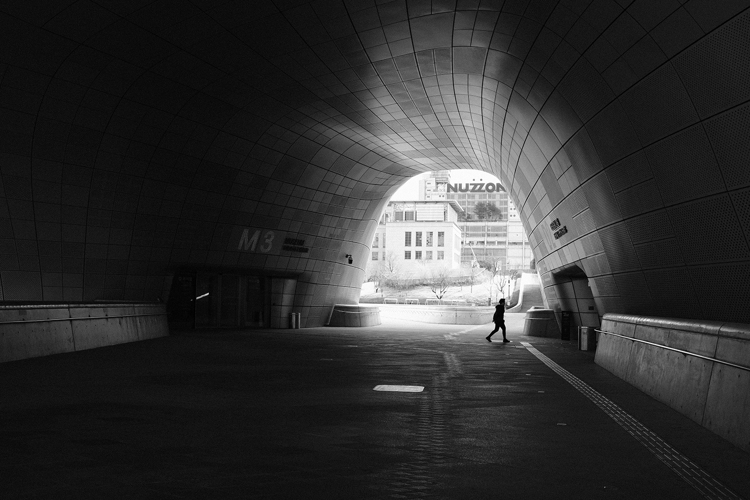 A lone figure walking toward the bright opening of a curved, tiled tunnel at Dongdaemun Design Plaza
