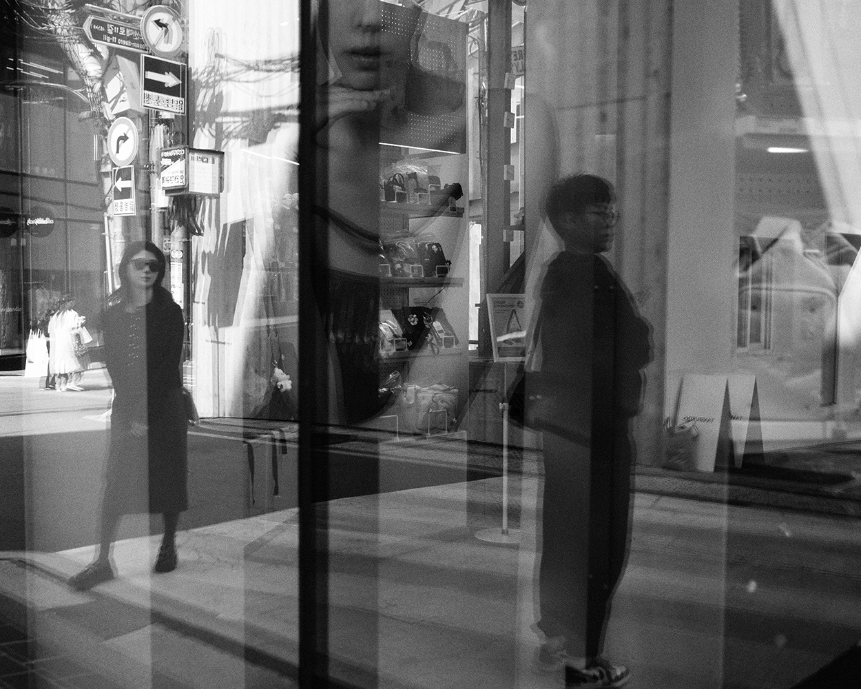 Layered shop-window reflections of a woman in sunglasses and a young man standing on a Seoul shopping street, a large poster of a face behind them