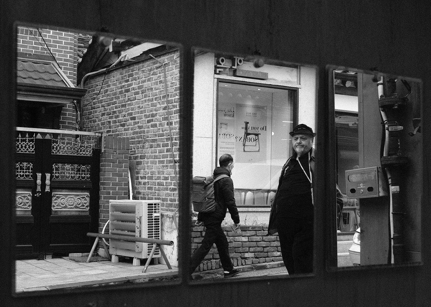 Three wall mirrors reflecting a brick alley, a masked man with a backpack walking past, and an older bearded man in a cap looking toward the camera