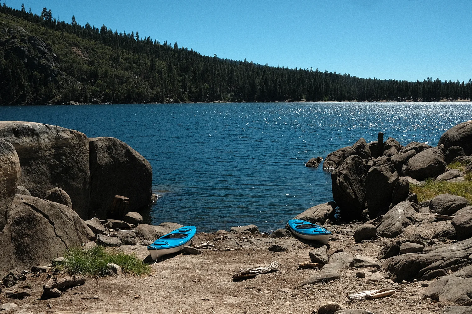 Two blue kayaks resting on a rocky inlet between large boulders, sunlight glittering on the lake