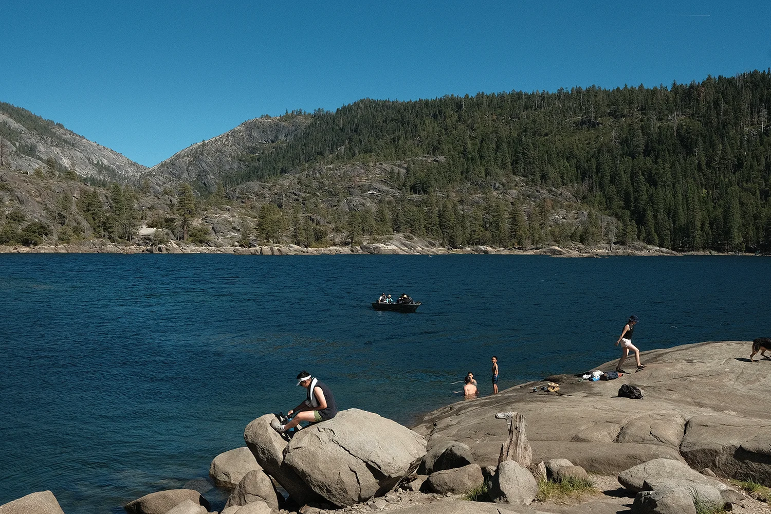 A man tying his shoes on a boulder by the lake while swimmers wade nearby and a small boat crosses the water