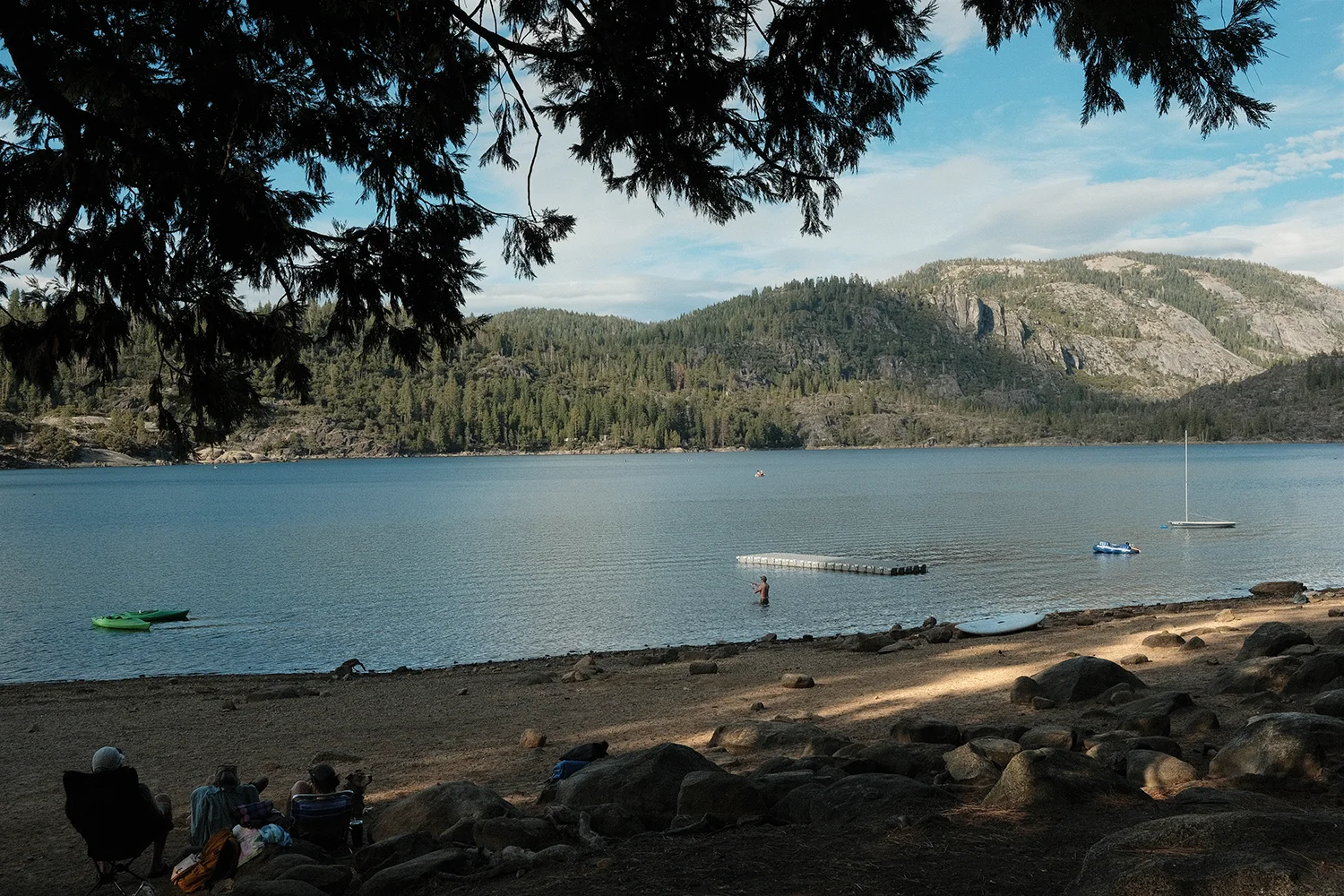 The lake framed by dark overhanging branches, with a swimmer, a floating dock, and a sailboat on the water