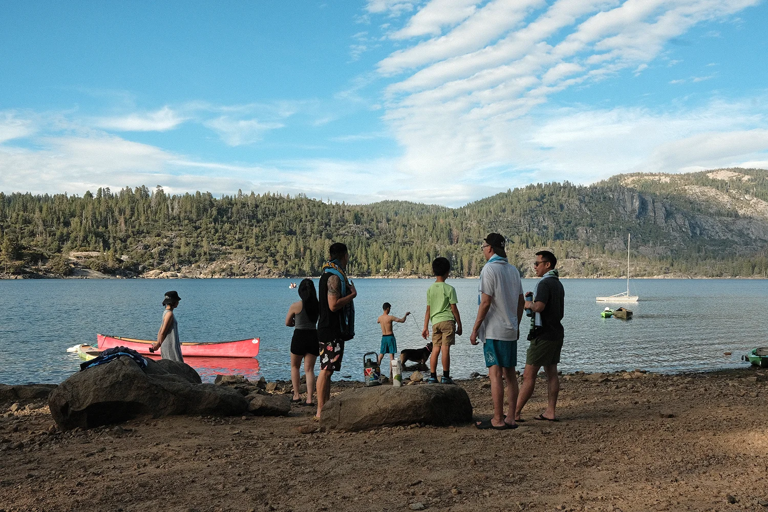 Friends and kids standing on the rocky shore at golden hour, a red canoe and a dog at the water’s edge